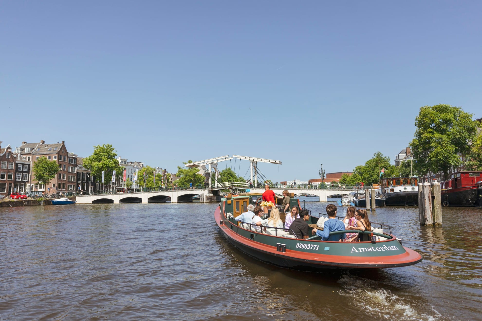 A classic cruise boat full of passengers sails along an Amsterdam canal towards the Magere Brug on a sunny day.