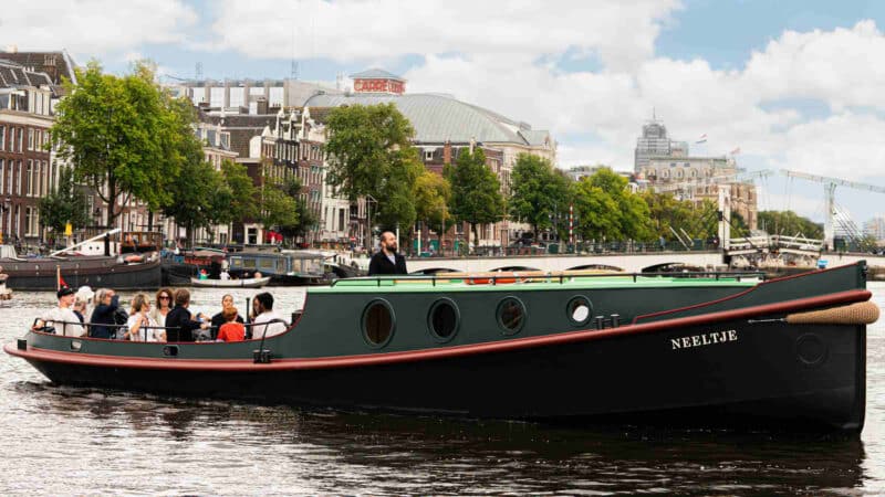 A tour boat with passengers sails under a stone arch bridge on the Amsterdam canals, while pedestrians walk across the bridge.